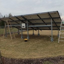 S.K.I. Master twoja złota rączka Kamil Dolata - Montaż paneli fotowoltaicznych na konstrukcji wsporczej, widok z dołu, drabiny i skrzynka narzędziowa obok. Instalacja w trakcie realizacji na trawiastym terenie.