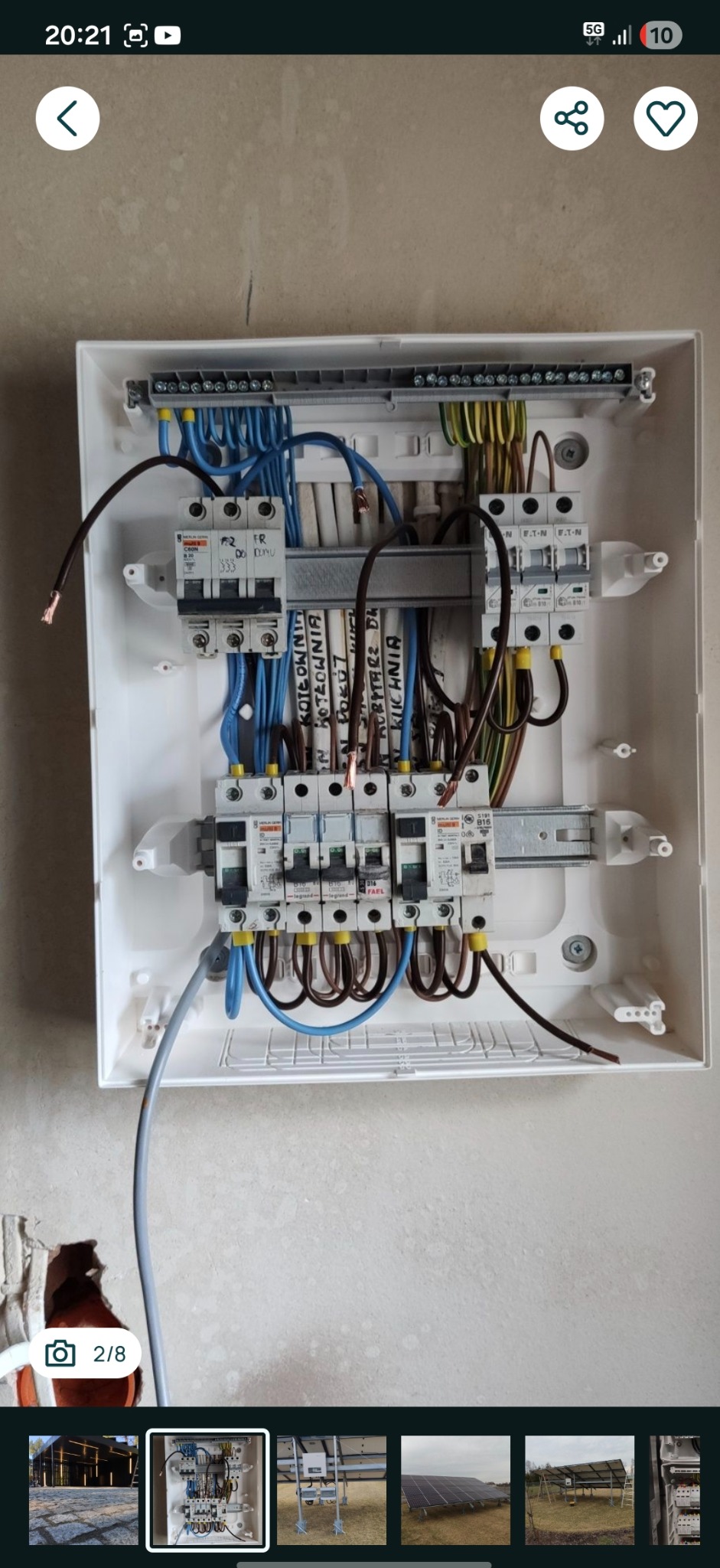 Rozdzielnica elektryczna w białej obudowie, z widocznymi bezpiecznikami, przewodami w kolorach niebieskim, brązowym i żółto-zielonym. Opisane obwody na listwie.