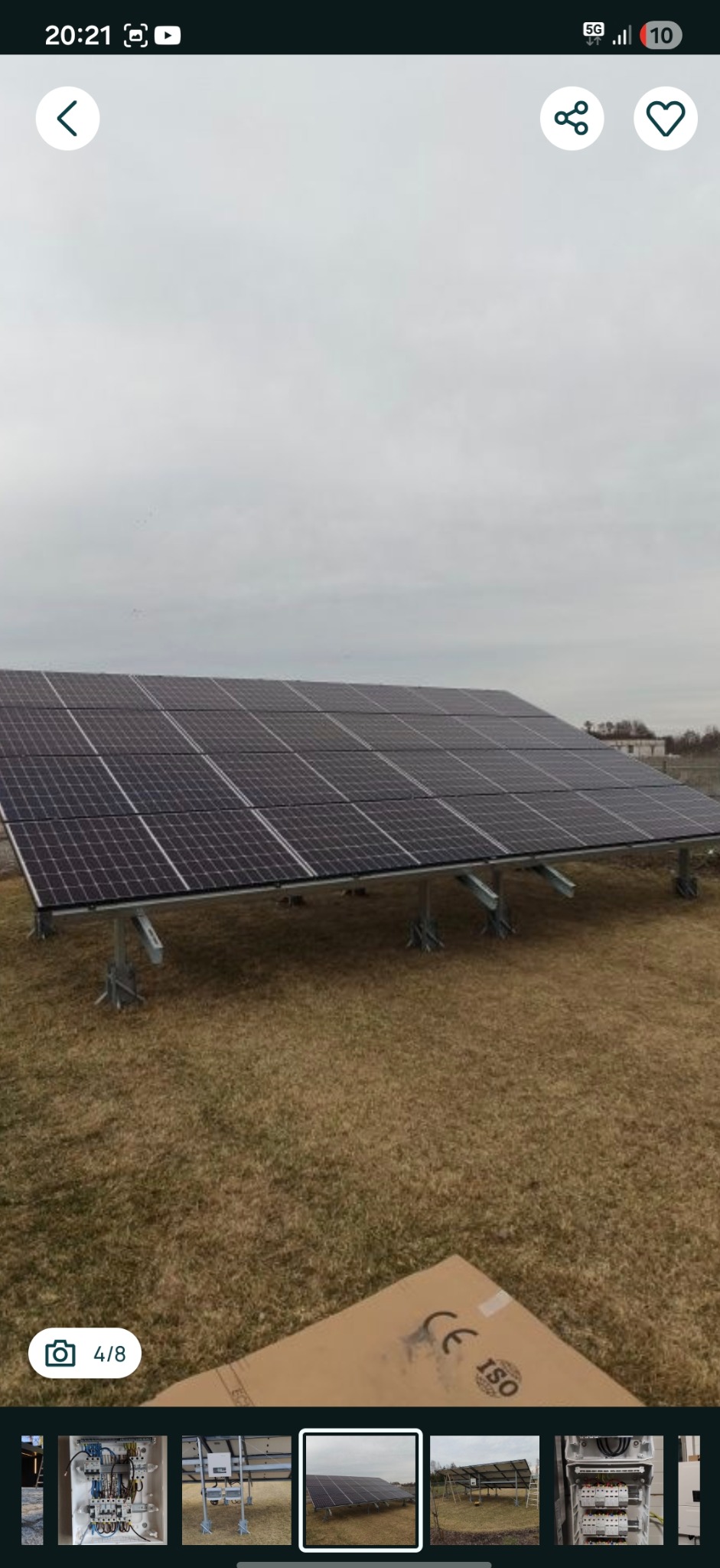 Panel słoneczny na trawiastym terenie, widoczne podpory i fragment kartonu z oznaczeniami CE i ISO. Pochmurne niebo w tle. Instalacja fotowoltaiczna.