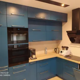 Niebieska zabudowa kuchenna z frontami na wysoki połysk, złotymi uchwytami, piekarnikiem Samsung, ekspresem do kawy i robotem kuchennym na drewnianym blacie.