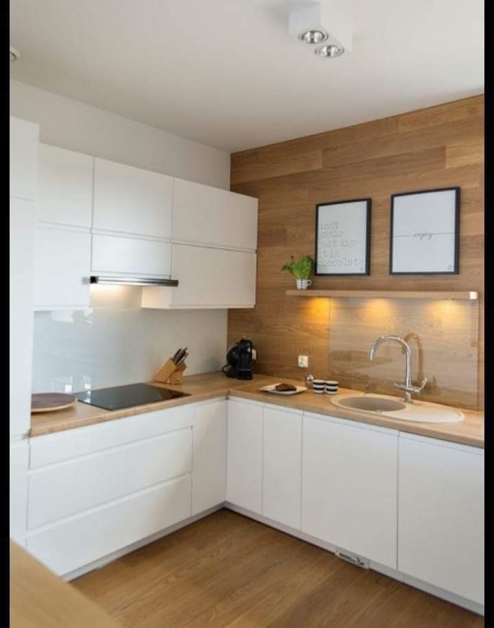 Jasna kuchnia z białymi frontami szafek, drewnianym blatem i panelami ściennymi, minimalistycznym oświetleniem i dekoracjami ściennymi, widok na narożnik i fragment podłogi z paneli.