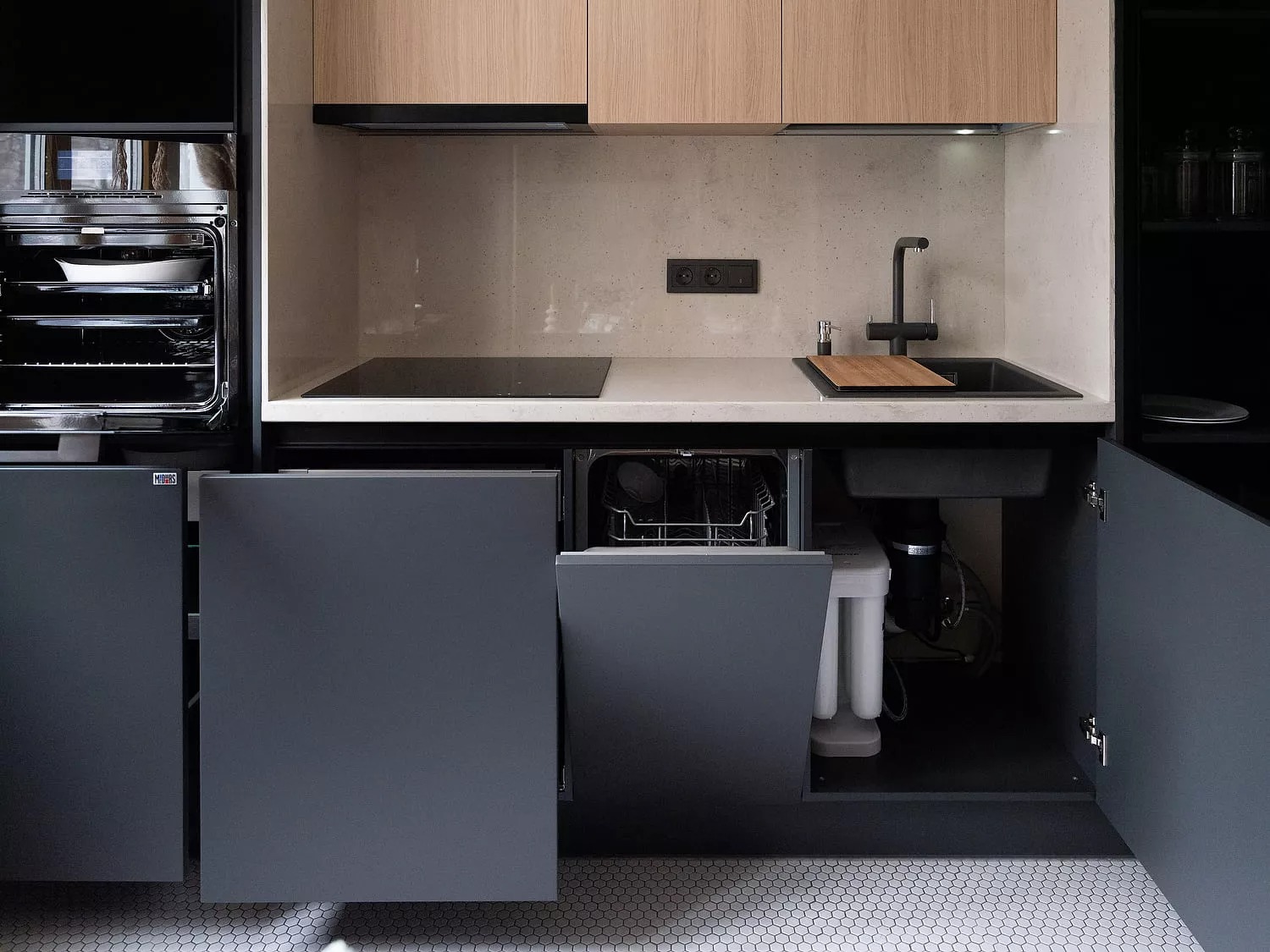 Nowoczesna kuchnia z szarymi frontami, drewnianymi górnymi szafkami i białym blatem. Widoczny piekarnik z otwartymi drzwiami, płyta indukcyjna, czarny zlew z baterią oraz otwarta zmywarka i szafka...