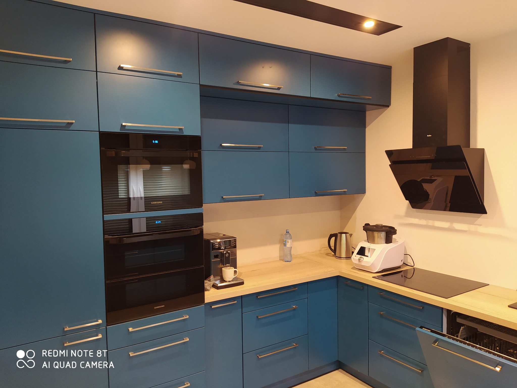 Niebieska zabudowa kuchenna z frontami na wysoki połysk, złotymi uchwytami, piekarnikiem Samsung, ekspresem do kawy i robotem kuchennym na drewnianym blacie.