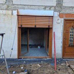 Spajder usługi montazowe - Montaż brązowych rolet zewnętrznych i okna w surowym stanie budynku, widoczne narzędzia i poziomica, częściowo opuszczona roleta.