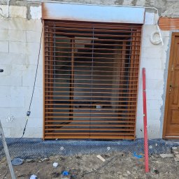 Spajder usługi montazowe - Montaż zewnętrznej rolety w kolorze brązowym na surowej ścianie z betonu komórkowego, widoczny poziomica, narzędzia i przewody na ziemi.