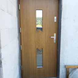 Zewnętrzne drzwi wejściowe w kolorze drewna z dwoma szybkami i wzorem, zamontowane w surowym otworze ściany budynku w trakcie prac wykończeniowych.