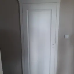 Białe drzwi wewnętrzne z ozdobną ramą i listwą przysufitową, widoczne na tle szarej ściany z włącznikiem światła.