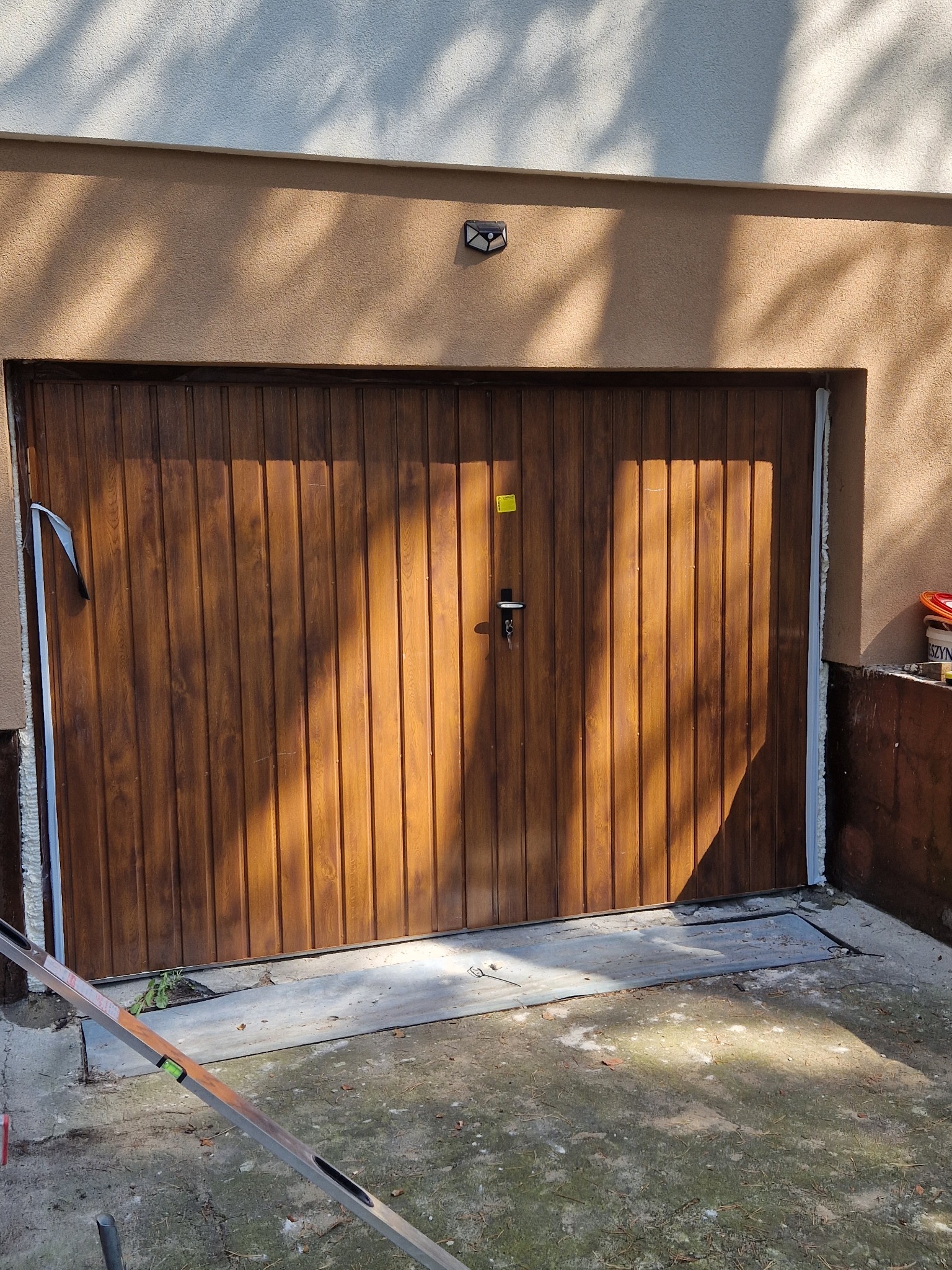 Montaż segmentowej bramy garażowej z drewnopodobnym wykończeniem; widoczne uszczelnienia, poziomica i cień rzucany przez drzewo.