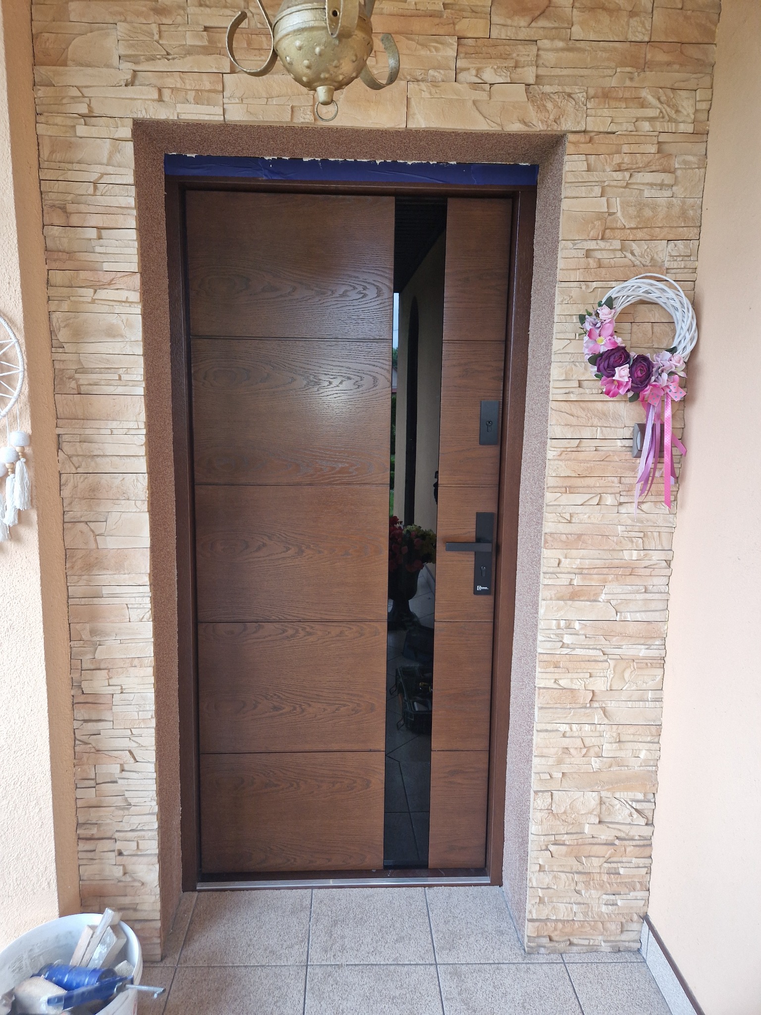 Nowoczesne drzwi zewnętrzne z poziomymi panelami w odcieniach brązu, zamontowane w Myszkowie, z dekoracyjną obudową z kamienia elewacyjnego i wieńcem kwiatowym po prawej stronie.