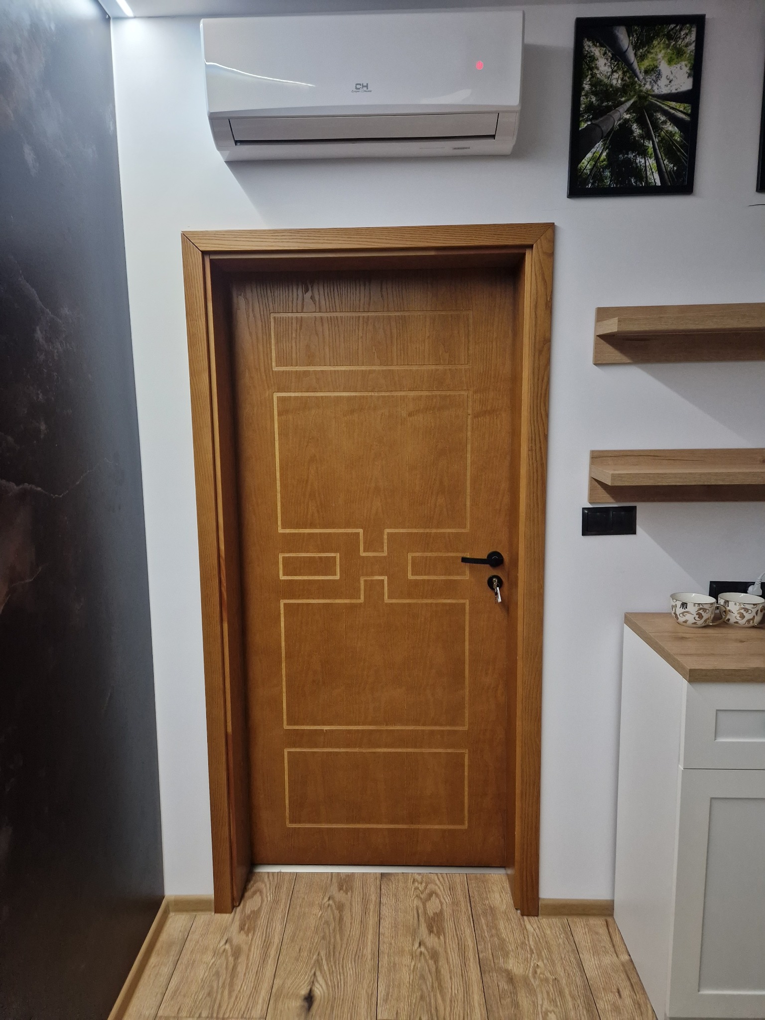 Dębowa stolarka drzwiowa wewnętrzna z geometrycznym zdobieniem, czarna klamka z kluczem, widok na klimatyzator, półki i fragment blatu kuchennego w jasnym wnętrzu.