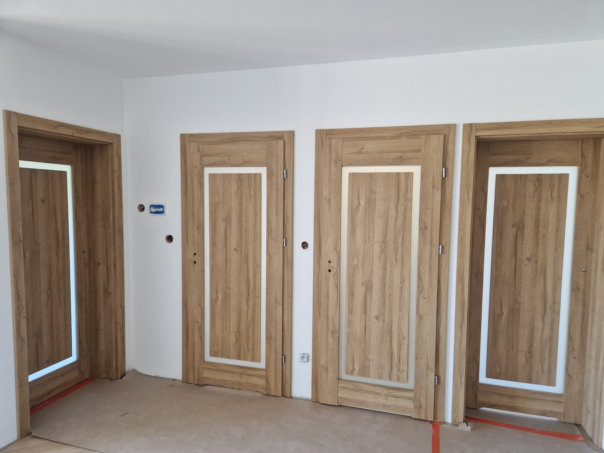 Montaż trzech drewnopodobnych drzwi wewnętrznych z białymi wstawkami w nowym, jeszcze nie wykończonym pomieszczeniu z widocznymi gniazdkami elektrycznymi i zabezpieczeniem podłogi.