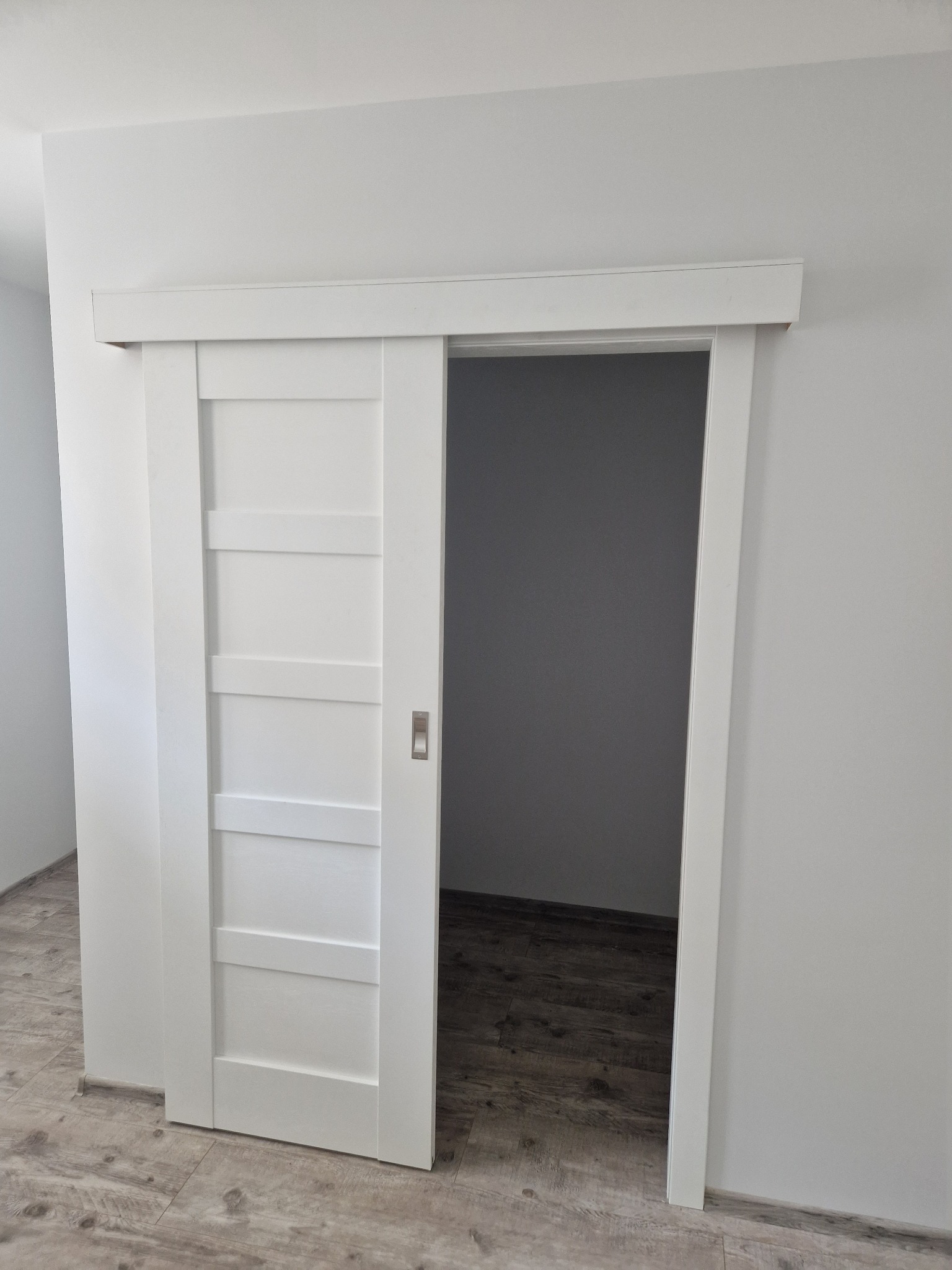 Białe, czteropanelowe drzwi przesuwne z metalowym zamkiem wpuszczanym, częściowo otwarte, ukazujące szare wnętrze pomieszczenia i drewnopodobną podłogę.