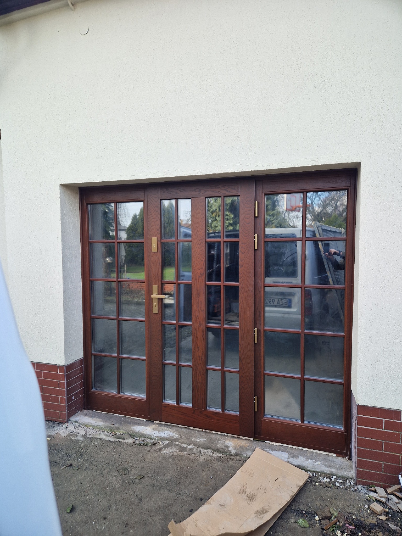 Zewnętrzne, drewniane, dwuskrzydłowe drzwi z wieloma szybkami, złote klamki, widoczne odbicie samochodu w szybach, surowy stan otoczenia.