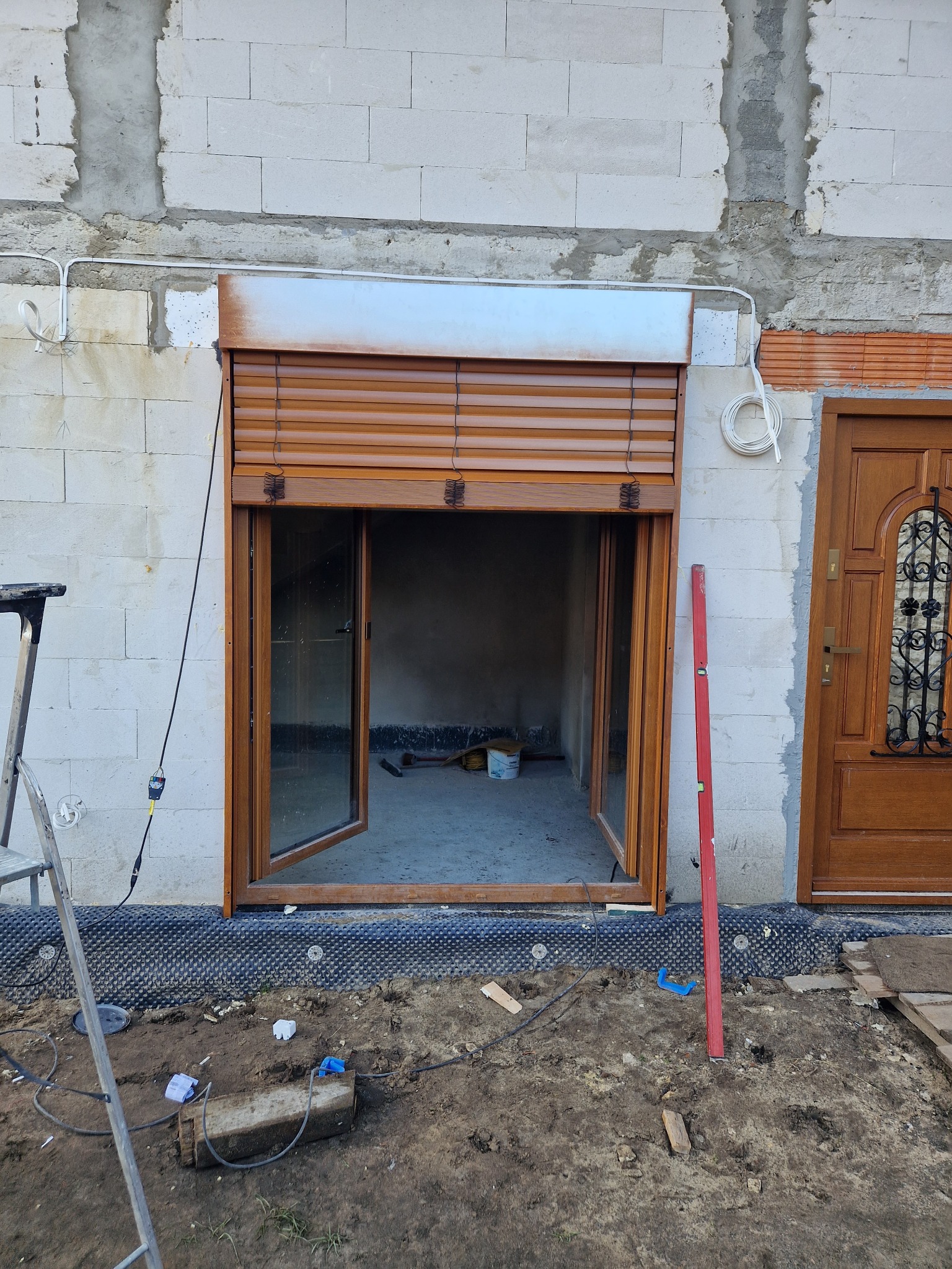 Montaż brązowych rolet zewnętrznych i okna w surowym stanie budynku, widoczne narzędzia i poziomica, częściowo opuszczona roleta.