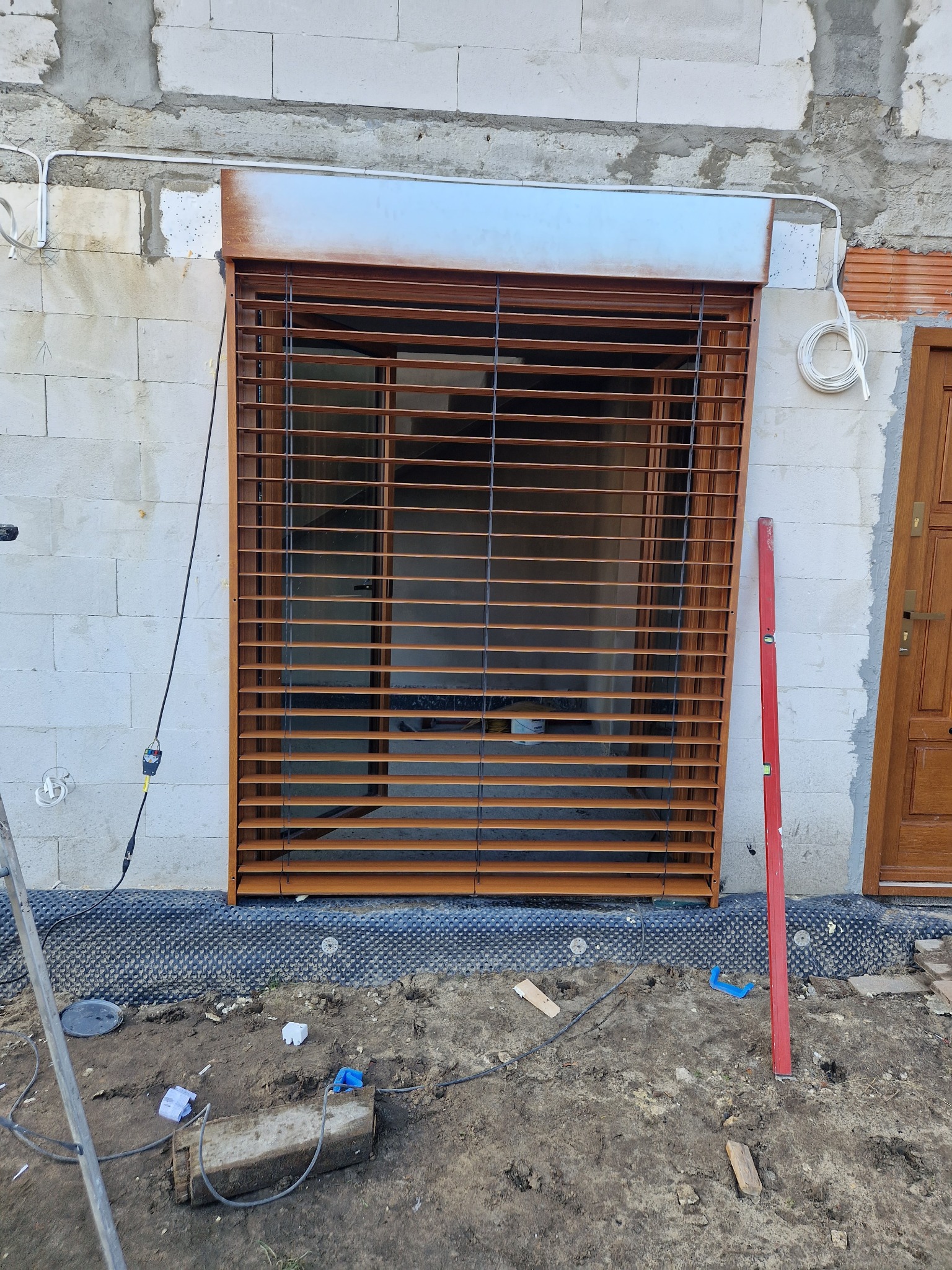 Montaż zewnętrznej rolety w kolorze brązowym na surowej ścianie z betonu komórkowego, widoczny poziomica, narzędzia i przewody na ziemi.