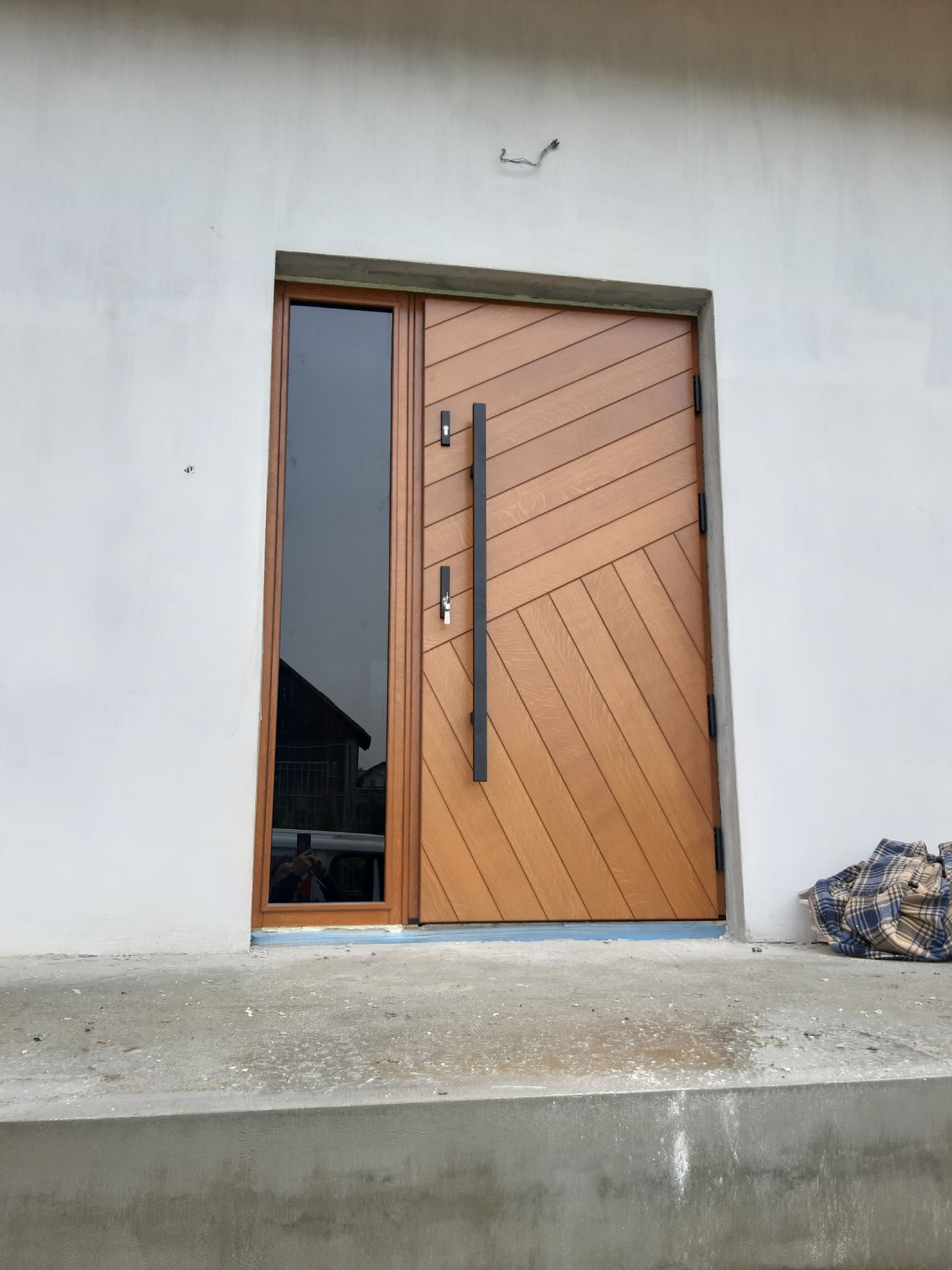 Nowoczesne drzwi wejściowe z drewna o skośnym wzorze, z wysokim, wąskim oknem po lewej stronie, zamontowane w surowym, białym murze.