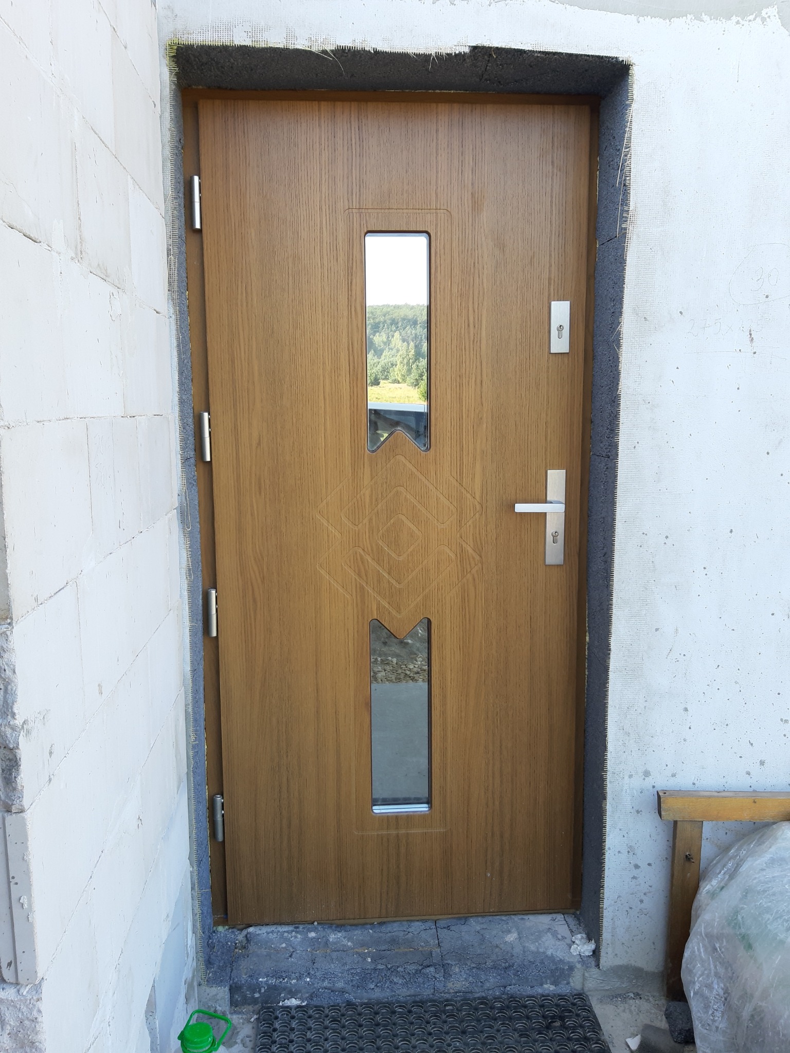 Zewnętrzne drzwi wejściowe w kolorze drewna z dwoma szybkami i wzorem, zamontowane w surowym otworze ściany budynku w trakcie prac wykończeniowych.