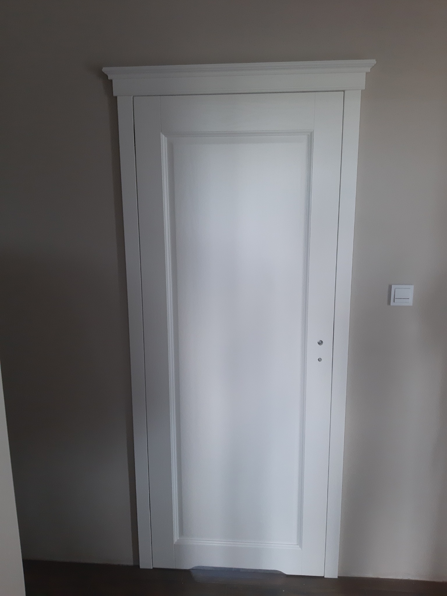 Białe drzwi wewnętrzne z ozdobną ramą i listwą przysufitową, widoczne na tle szarej ściany z włącznikiem światła.