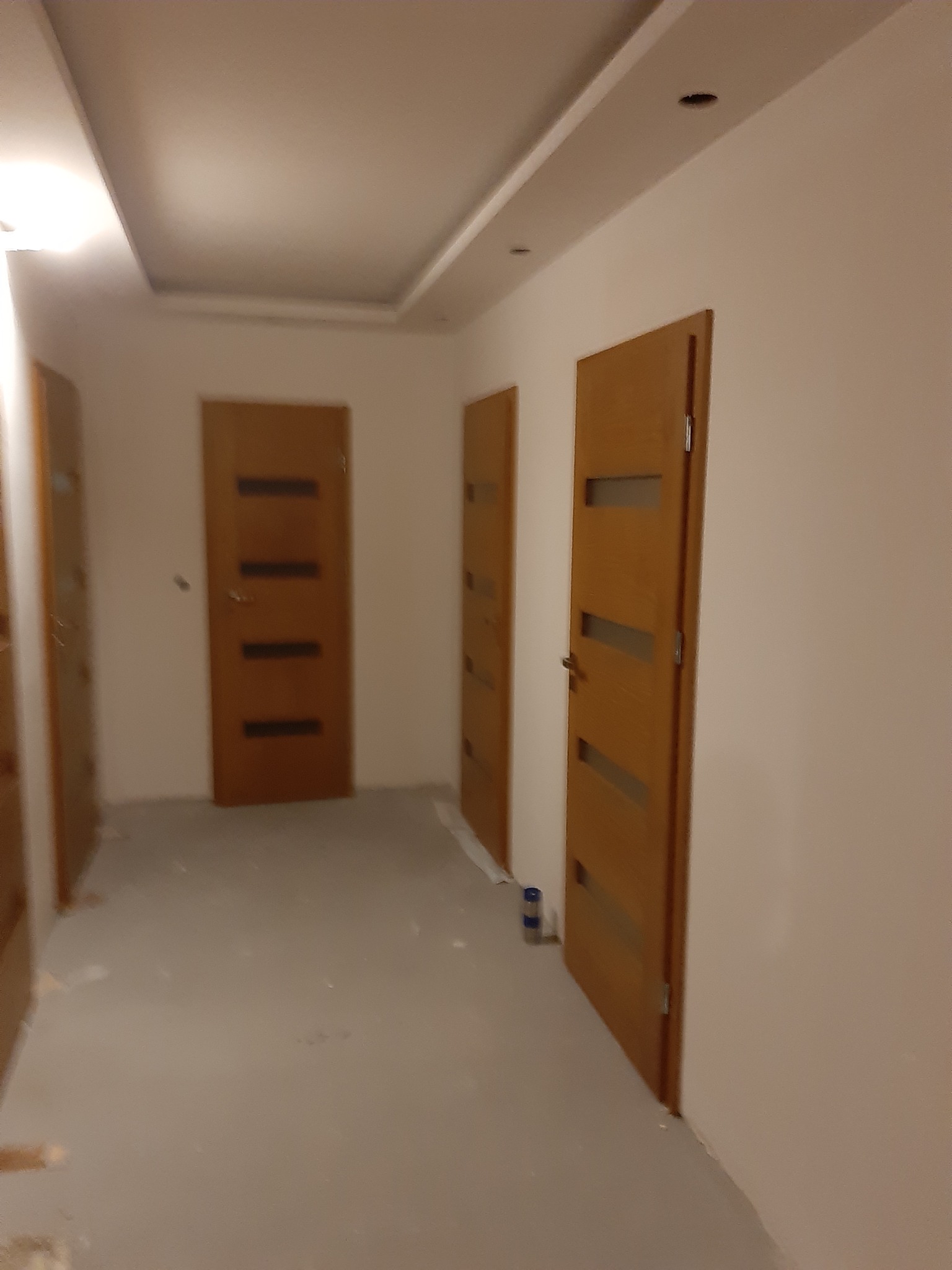 Wnętrze korytarza w trakcie remontu z trzema parami drewnianych drzwi wewnętrznych z poziomymi przeszkleniami, widoczne ślady po malowaniu na podłodze.