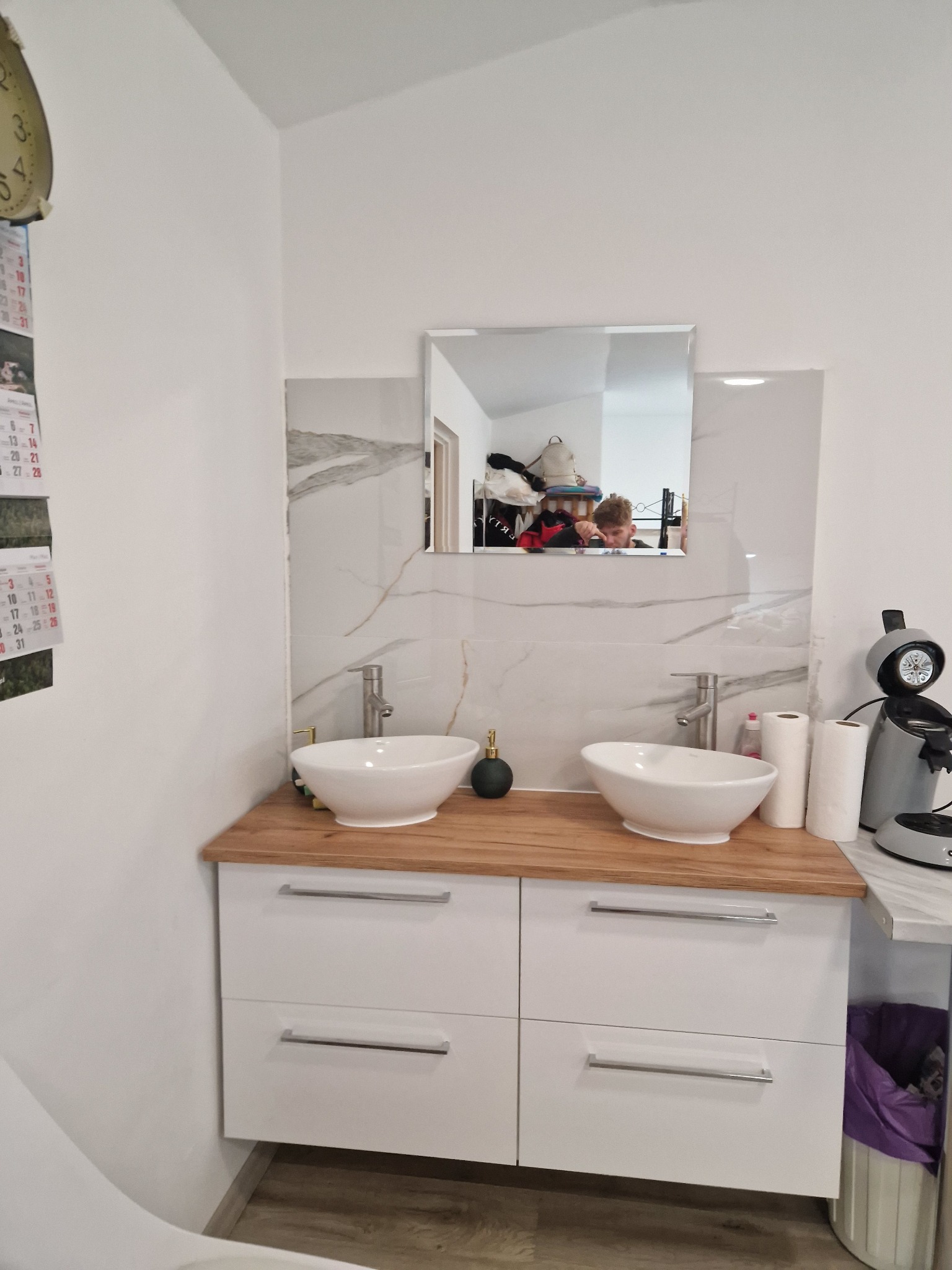 Nowoczesna łazienka z dwoma umywalkami nablatowymi w białych misach, drewnianym blatem i białymi szafkami z szufladami, wykończona płytkami imitującymi marmur. W lustrze odbija się osoba robiąca...