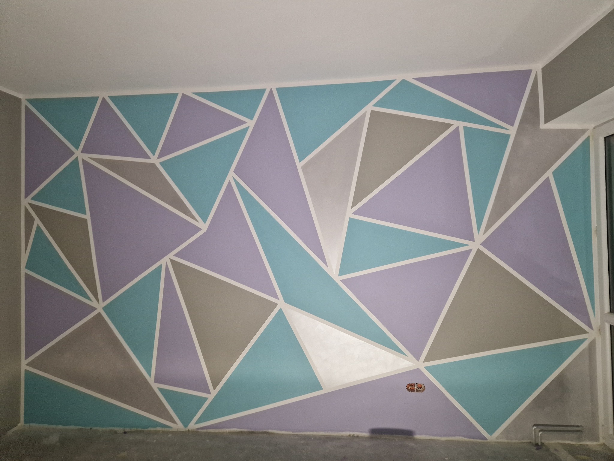 Ściana w pokoju pomalowana w geometryczne wzory: trójkąty w odcieniach fioletu, turkusu i szarości, oddzielone białymi liniami. Widoczna puszka elektryczna w dolnej części ściany.