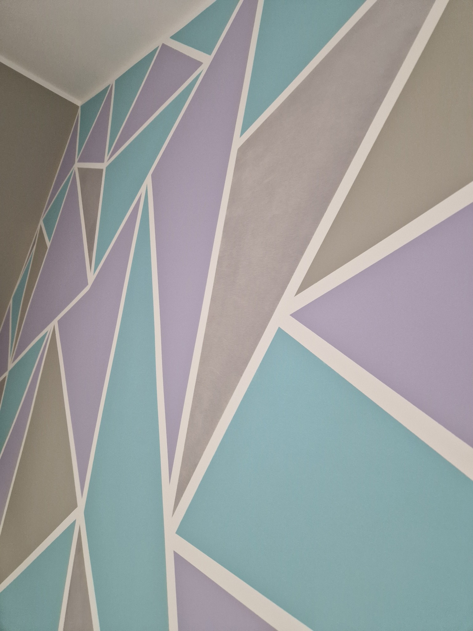 Ściana z geometrycznym wzorem w pastelowych odcieniach błękitu, fioletu i szarości, oddzielonych białymi liniami, widziana pod kątem w rogu pomieszczenia.