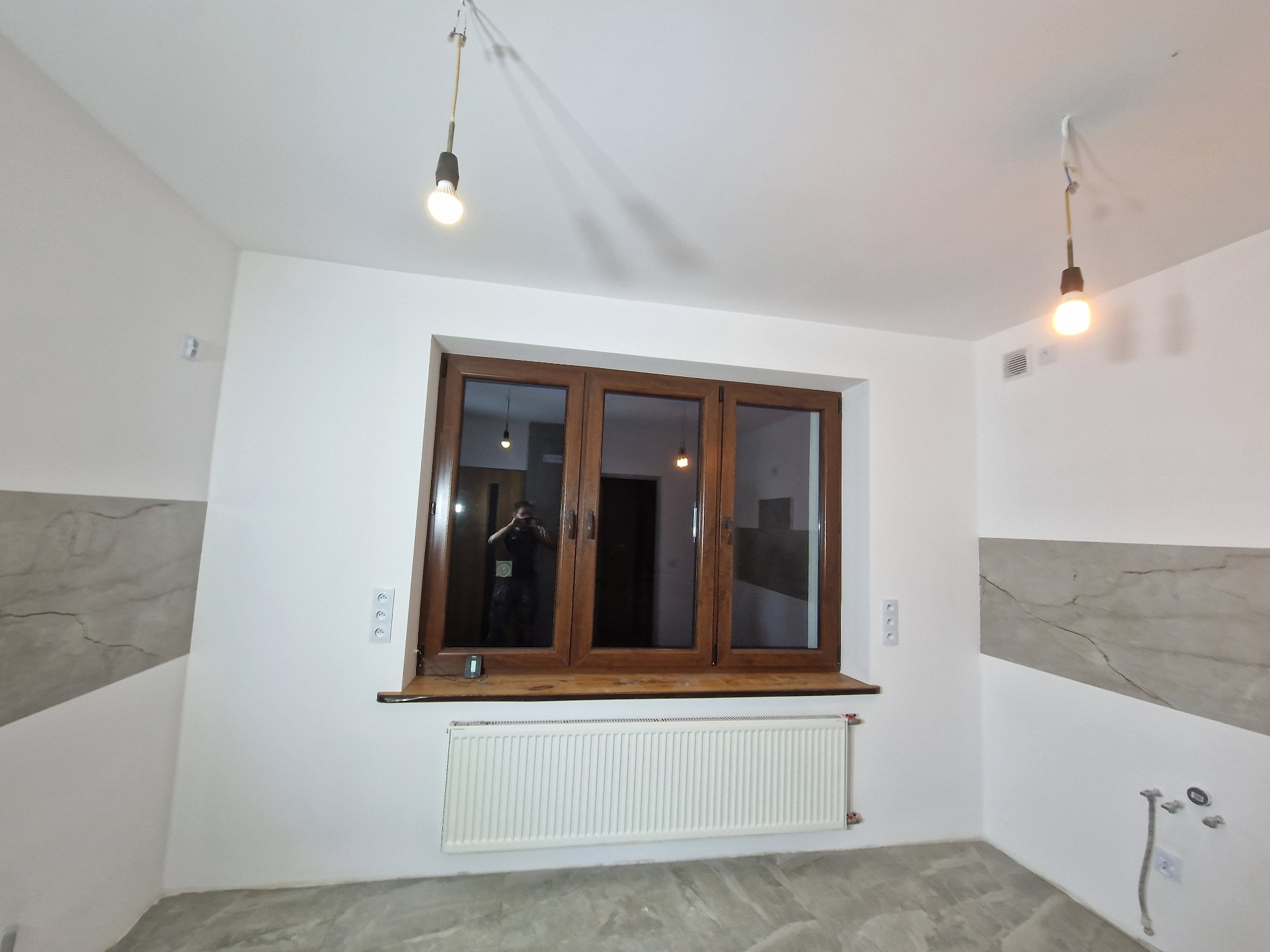 Wykończone wnętrze pokoju z drewnianą stolarką okienną, białymi ścianami z szarym akcentem imitującym marmur, grzejnikiem pod oknem i betonową wylewką na podłodze, oświetlone dwoma wiszącymi...