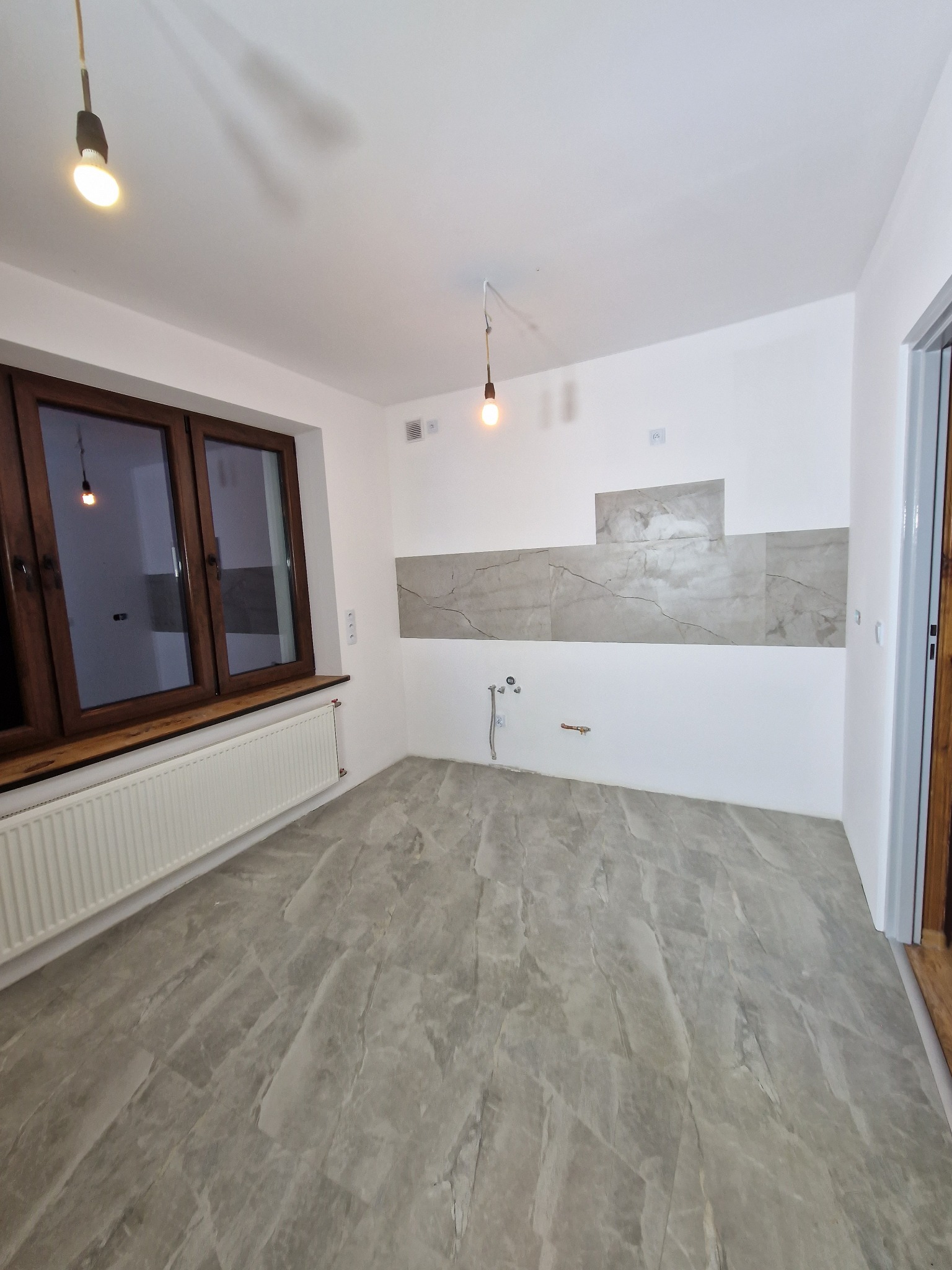 Wnętrze kuchni w trakcie prac wykończeniowych: położona szara podłoga imitująca marmur, częściowo wykończona ściana nad blatem z szarymi panelami, widoczne instalacje wodne, drewniane okno i grzejnik.