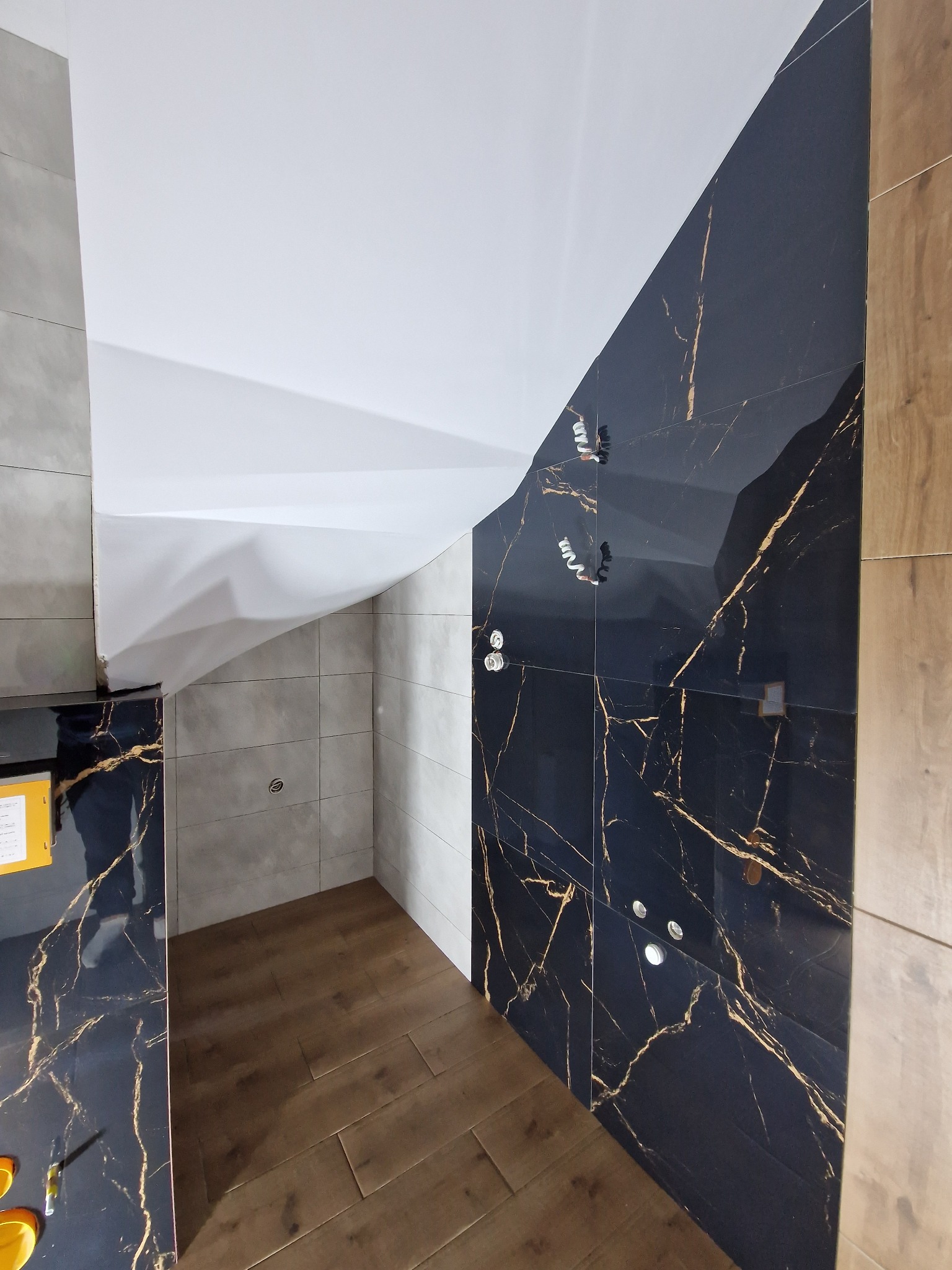 Wykończona łazienka z czarnymi płytkami imitującymi marmur ze złotymi żyłami, drewnopodobną podłogą i podwieszanym sufitem z oświetleniem punktowym.