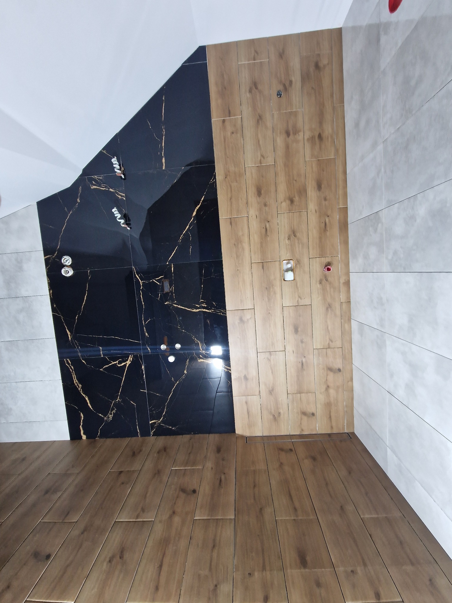 Wykończona łazienka z czarną marmurową ścianą, drewnopodobnymi płytkami i podłogą z paneli, widoczne instalacje elektryczne.