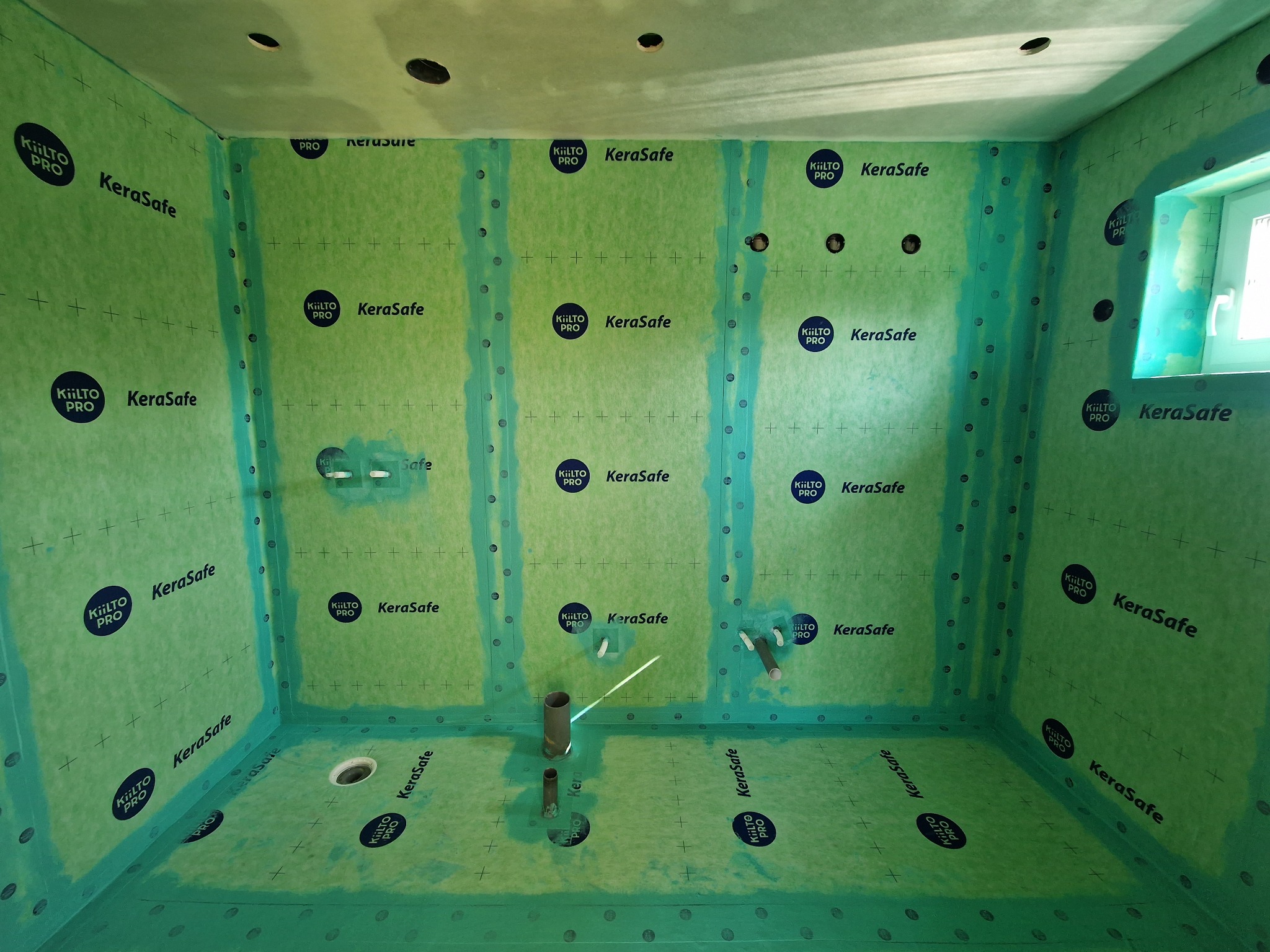 Przygotowana do hydroizolacji łazienka z zieloną matą KeraSafe, widoczne wyprowadzenia instalacji wodnej i kanalizacyjnej oraz otwory w suficie na oświetlenie.