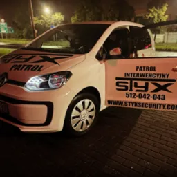 Biały samochód patrolowy z otwartymi drzwiami, logo firmy 'Styx' i numerem telefonu widoczne na karoserii, nocne oświetlenie uliczne w tle.