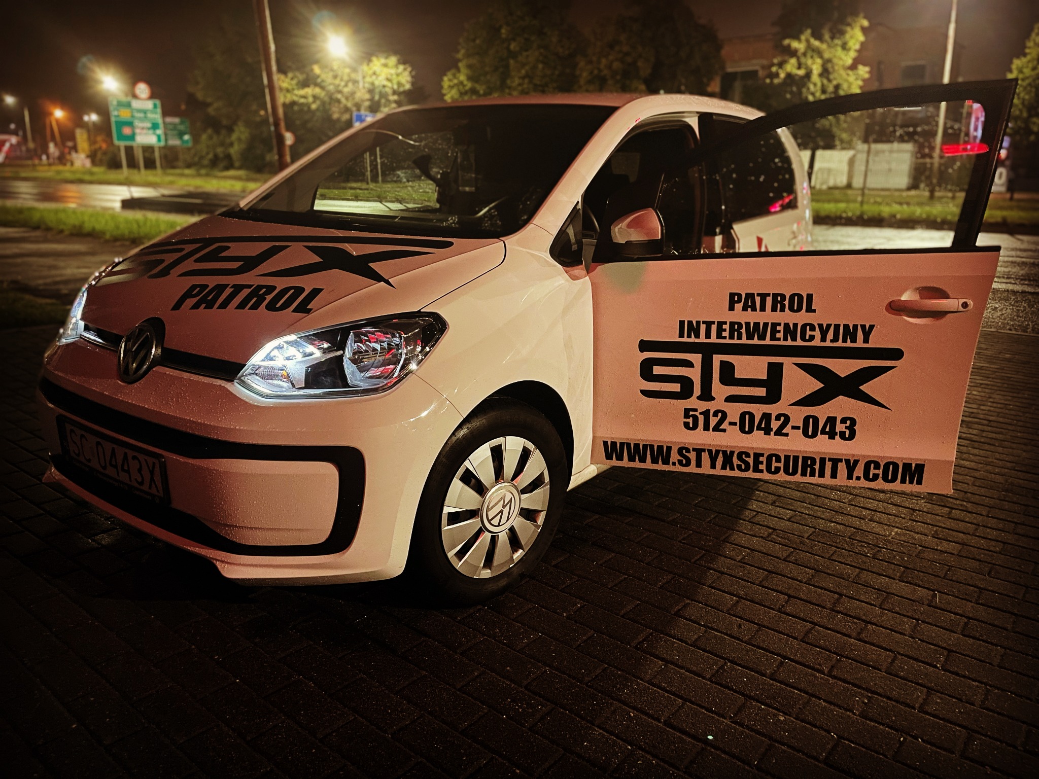 Biały samochód patrolowy z otwartymi drzwiami, logo firmy 'Styx' i numerem telefonu widoczne na karoserii, nocne oświetlenie uliczne w tle.