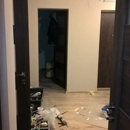 BAD-BUD - Widok przez otwarte drzwi na pokój w trakcie remontu; widoczne narzędzia, kartony, wiadro i odkurzacz przemysłowy; ściany przygotowane pod malowanie.
