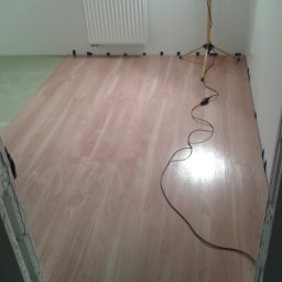 BAD-BUD - Nowa podłoga laminowana w trakcie montażu, zabezpieczona niebieską taśmą malarską przy ścianach, widoczna lampa robocza i kabel zasilający.