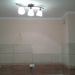 BAD-BUD - Wnętrze kuchni w trakcie remontu: częściowo wyłożone płytkami ściany, widoczne przewody elektryczne, sufit z lampą.