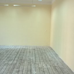 BAD-BUD - Pusta, jasna przestrzeń z pomalowanymi na kremowo ścianami i starym, drewnianym parkietem wymagającym renowacji, oświetlona przez podłużne lampy sufitowe.