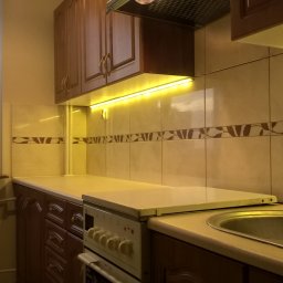 BAD-BUD - Wykończona kuchnia z brązowymi szafkami, beżowymi płytkami z ciemnobrązowym wzorem i oświetleniem LED pod szafkami, z widocznym oknem i fragmentem dłoni w lewym górnym rogu.