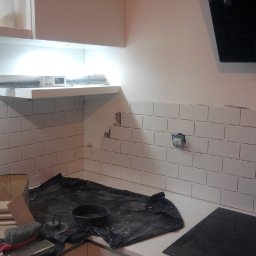 BAD-BUD - Białe, prostokątne płytki ułożone na ścianie w kuchni, widoczne narzędzia i materiały budowlane, fragment niezabezpieczonego gniazdka elektrycznego.