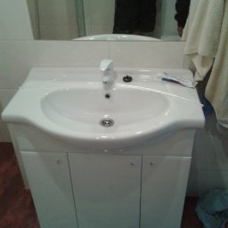 BAD-BUD - Biała umywalka z szafką w łazience, widoczna tuba pasty do zębów, odbicie osoby w lustrze, ręcznik i ubrania na podłodze.