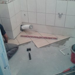 BAD-BUD - Układanie beżowych płytek podłogowych w łazience, widoczny fragment instalacji kanalizacyjnej, poziomica, młotek i wiadro z zaprawą.