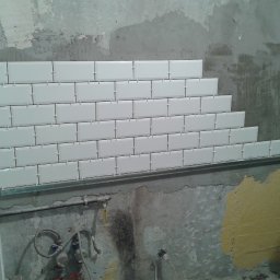 BAD-BUD - Układanie białych, prostokątnych płytek na ścianie z widocznymi rurami i fragmentami starej farby, użycie krzyżyków dystansowych.