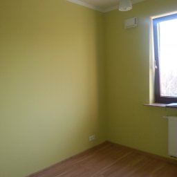 BAD-BUD - Pomalowane na oliwkowy kolor wnętrze pokoju z drewnianą podłogą, widoczne okno z brązową ramą, biały grzejnik i czarny żyrandol z białymi kloszami.