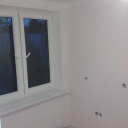 BAD-BUD - Pokój po malowaniu na jasny róż, widoczne gniazdka elektryczne bez osprzętu i okno z białymi ramami; podłoga w trakcie przygotowania.