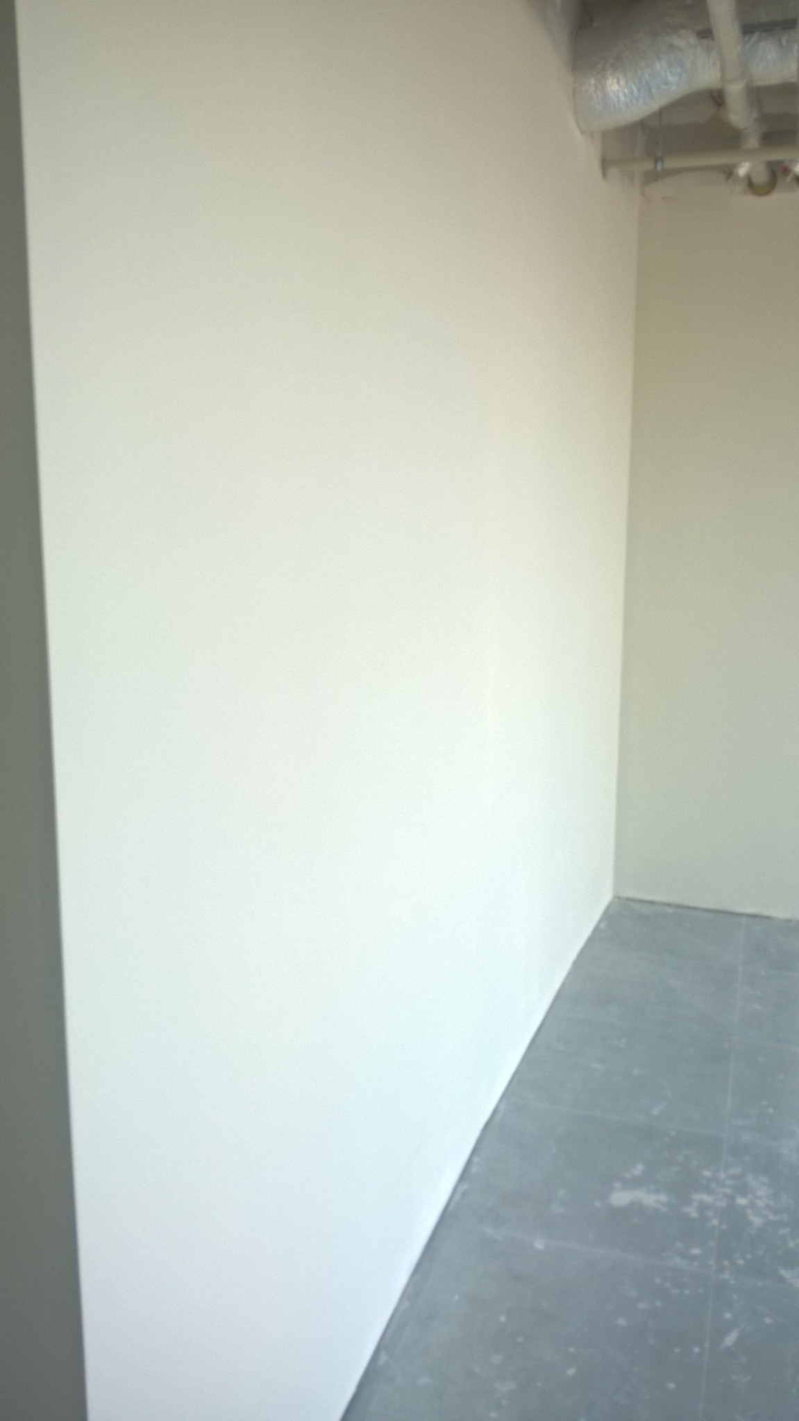 Pomieszczenie po malowaniu ścian na biało, widoczne szare płytki na podłodze i rury wentylacyjne pod sufitem.