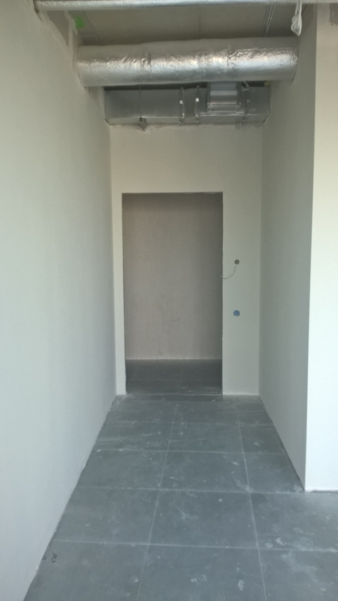 Wnętrze w trakcie wykańczania: szare płytki podłogowe, białe ściany, widoczny kanał wentylacyjny pod sufitem, otwór drzwiowy prowadzący do kolejnego pomieszczenia w stanie surowym.