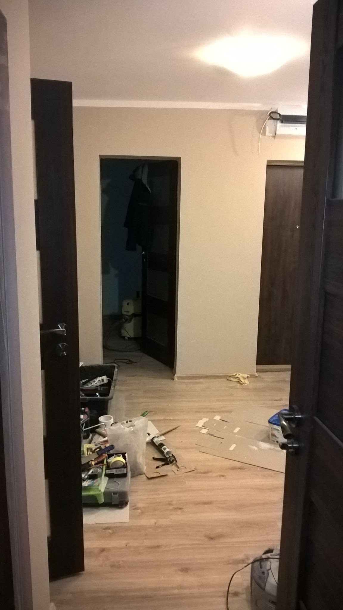 Widok przez otwarte drzwi na pokój w trakcie remontu; widoczne narzędzia, kartony, wiadro i odkurzacz przemysłowy; ściany przygotowane pod malowanie.