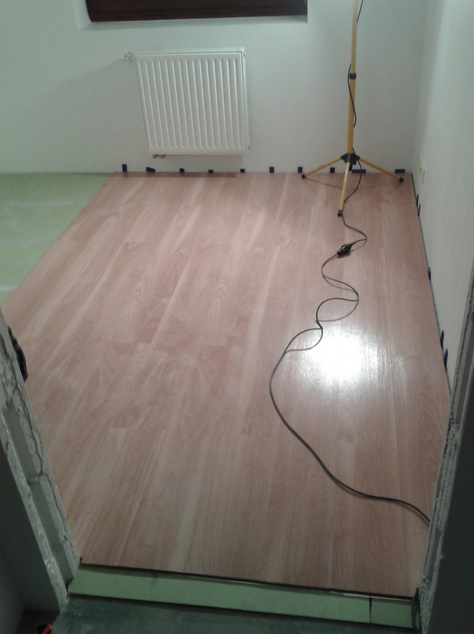 Nowa podłoga laminowana w trakcie montażu, zabezpieczona niebieską taśmą malarską przy ścianach, widoczna lampa robocza i kabel zasilający.