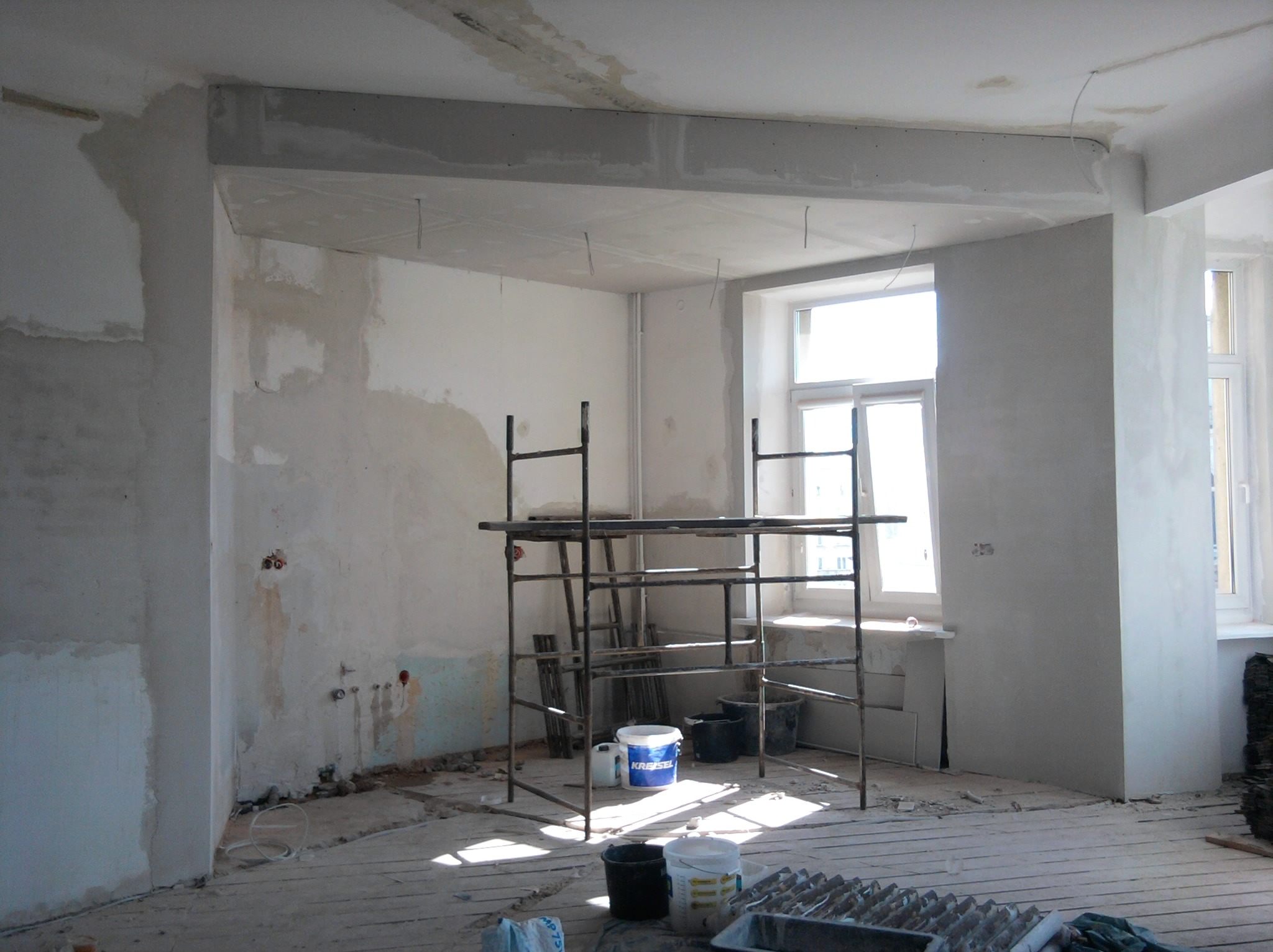 Pokój w trakcie prac wykończeniowych: surowe ściany z widocznymi śladami szpachlowania, metalowe rusztowanie pośrodku, wiadra z farbą i gładzią na podłodze, widoczne instalacje elektryczne...