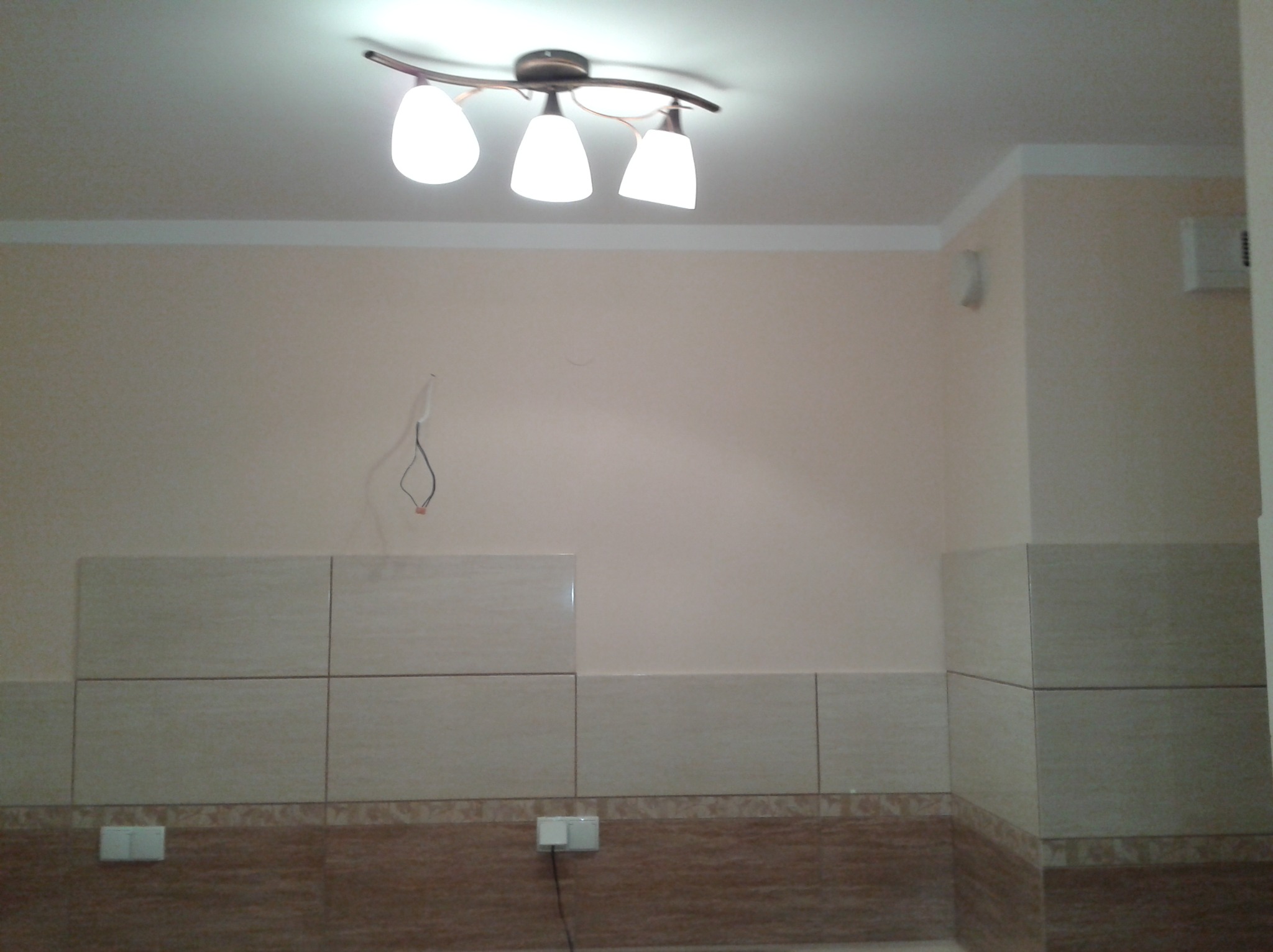 Wnętrze kuchni w trakcie remontu: częściowo wyłożone płytkami ściany, widoczne przewody elektryczne, sufit z lampą.